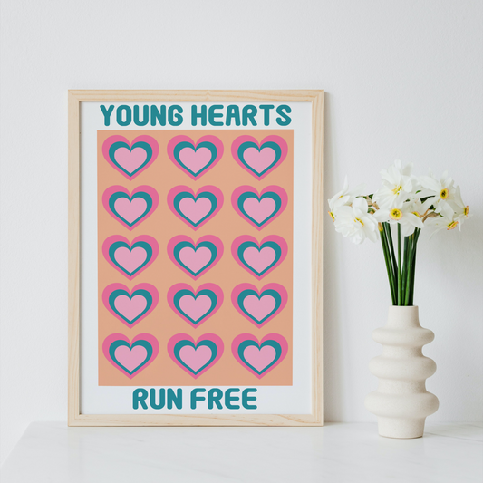 A framed art print with the text 'YOUNG HEARTS RUN FREE' and a grid of pink and teal hearts on a peach background, styled on a white surface next to a vase of white flowers against a white wall.