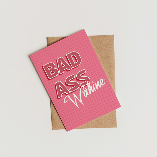 A pink grid-patterned greeting card with the words 'BAD ASS Wahine' in raised pink and white lettering, resting on top of a brown kraft paper envelope against a clean white background.