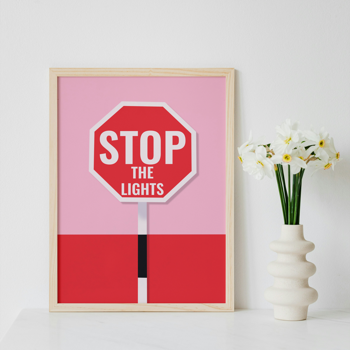 A framed art print featuring a stylized stop sign with the text 'STOP THE LIGHTS' on a pink and red background, displayed on a white surface next to a white vase with white flowers against a white wall.