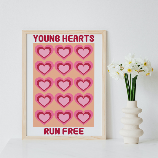 A framed art print with a light wood frame sits on a white surface next to a white ceramic vase holding white daffodils. The print features a grid of pink hearts on a peach background with the text 'YOUNG HEARTS' at the top and 'RUN FREE' at the bottom.