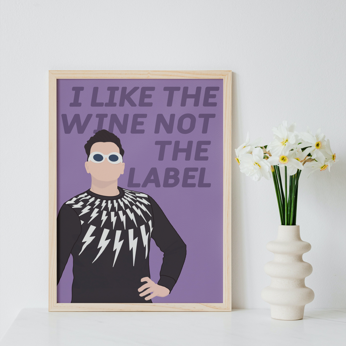 A framed art print featuring a stylized illustration of a man with the text 'I LIKE THE WINE NOT THE LABEL' on a purple background, displayed on a white surface next to a vase of white flowers against a light gray wall.