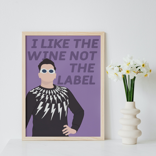 A framed art print featuring a stylized illustration of a man with the text 'I LIKE THE WINE NOT THE LABEL' on a purple background, displayed on a white surface next to a vase of white flowers against a light gray wall.