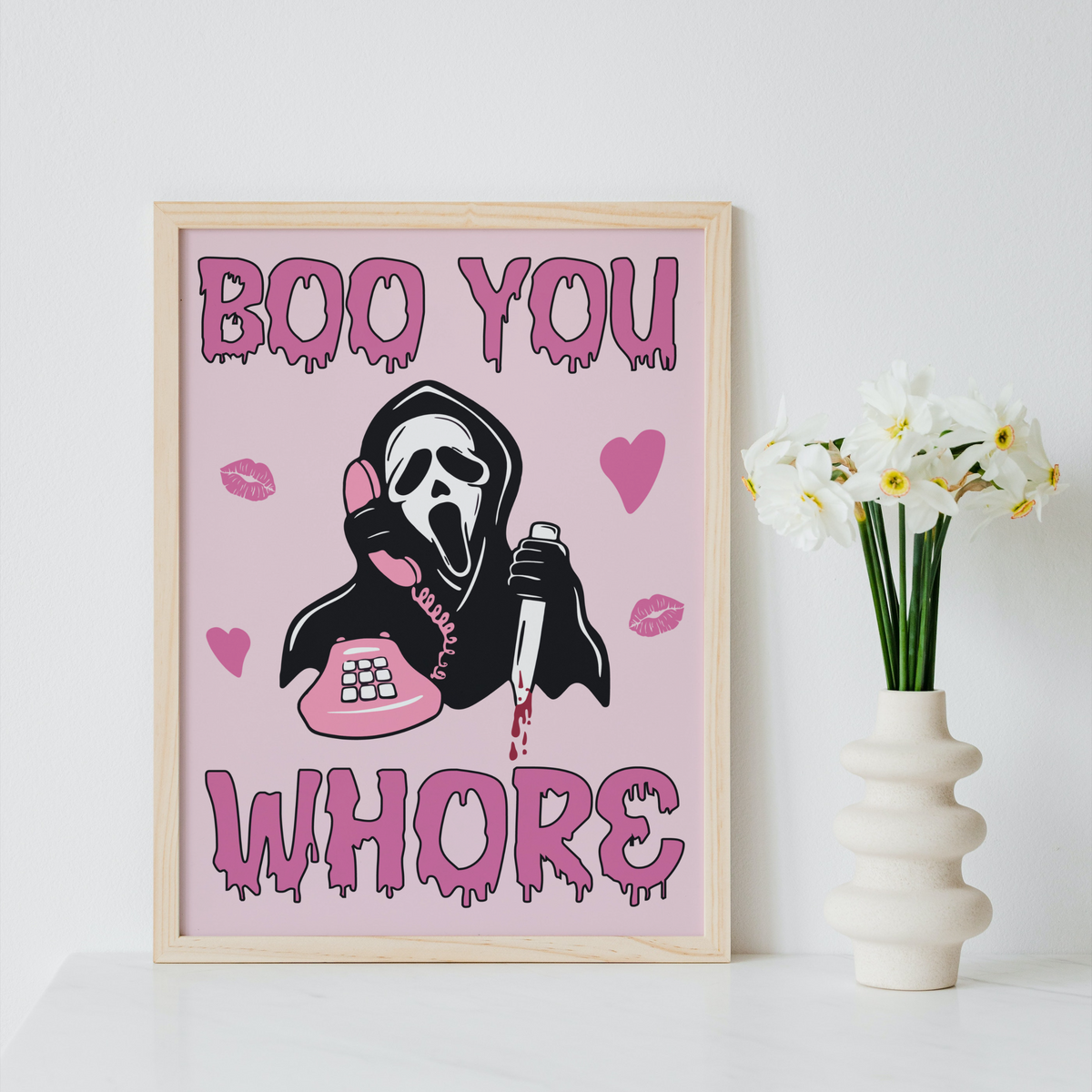 A framed art print featuring a black and white Ghostface figure holding a pink phone and blood, with the text 'BOO YOU WHORE' in dripping pink letters on a light pink background. The frame sits on a white surface next to a white vase with white flowers against a white wall.