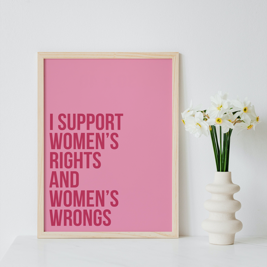A framed pink typographic art print with the text 'I SUPPORT WOMEN'S RIGHTS AND WOMEN'S WRONGS' in darker pink lettering, leaning against a white wall beside a white vase with white flowers.