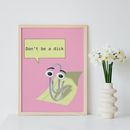 A framed novelty art print featuring a cartoon paperclip with eyes and a speech bubble that reads 'Don't be a dick' on a pink background, styled on a white surface next to a vase of white flowers.