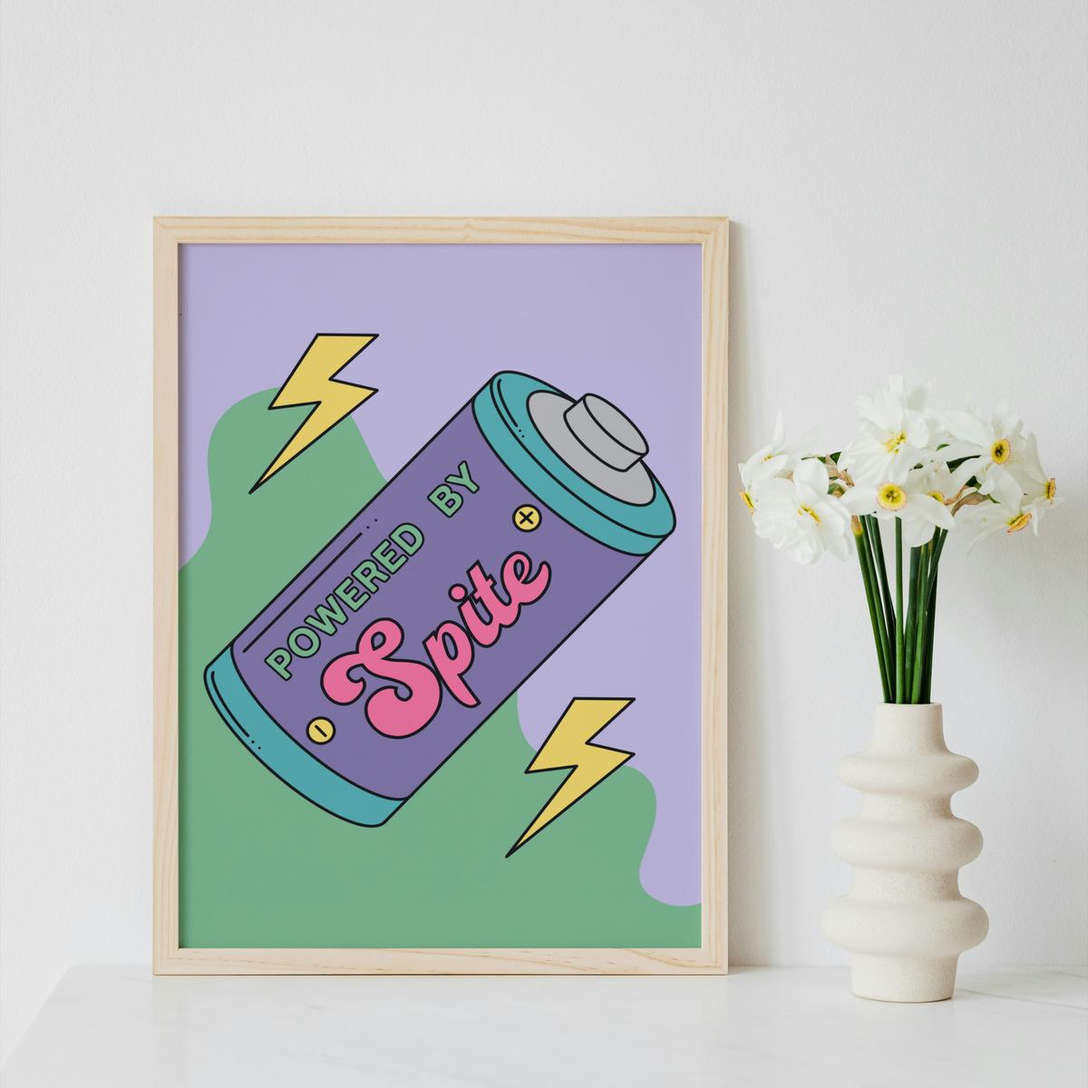 A framed art print featuring a retro-style spray can illustration with the words 'POWERED BY Spite' on a lavender and green background, styled on a white surface next to a white vase with white flowers against a white wall.