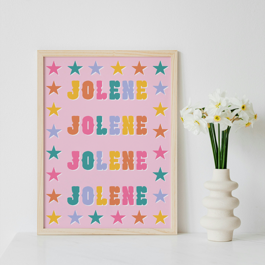 A framed art print featuring the name 'JOLENE' repeated in colorful letters on a pink background with pastel stars, styled on a white surface next to a white vase with white flowers against a white wall.