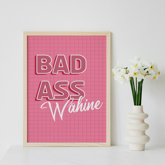 A framed typographic wall art print with the words “BAD ASS Wahine” on a pink grid background, leaning against a white wall beside a white vase with white flowers.