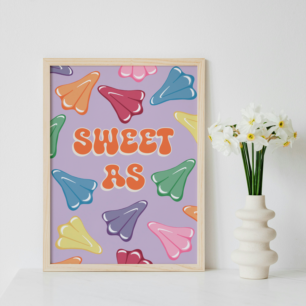 A framed art print featuring the text 'SWEET AS' in orange bubble letters over a lavender background with colorful candy-like shapes, styled on a white surface next to a white vase with white flowers.