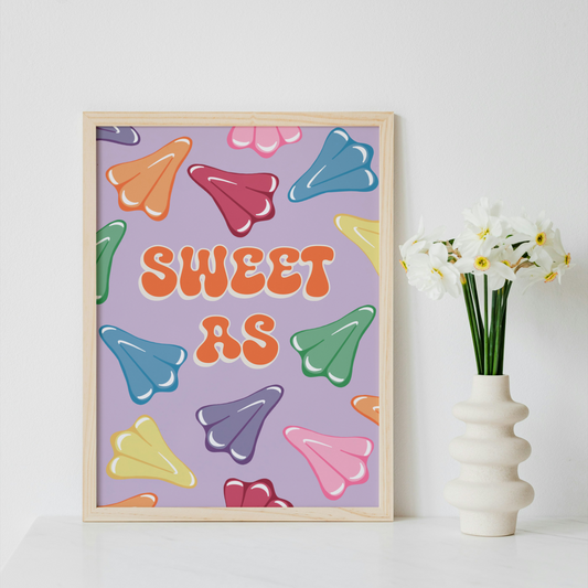 A framed art print featuring the text 'SWEET AS' in orange bubble letters over a lavender background with colorful candy-like shapes, styled on a white surface next to a white vase with white flowers.