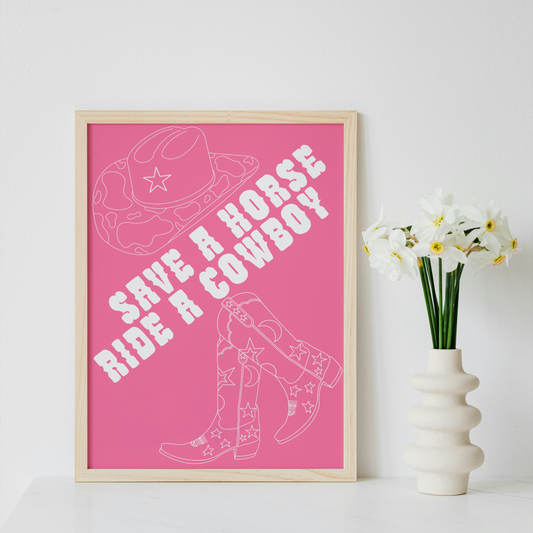A framed art print featuring a pink background with white western-themed line art and the text 'SAVE A HORSE RIDE A COWBOY', displayed on a white surface next to a white vase with white flowers against a white wall.