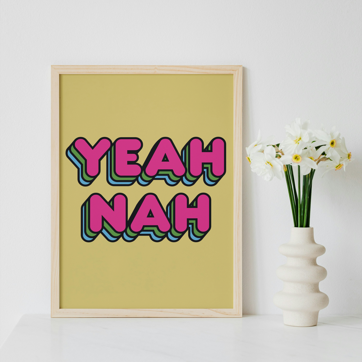 A framed typographic art print reading 'YEAH NAH' in pink bubble letters with blue and green shadowing, displayed on a white surface next to a white vase with white flowers against a white wall.