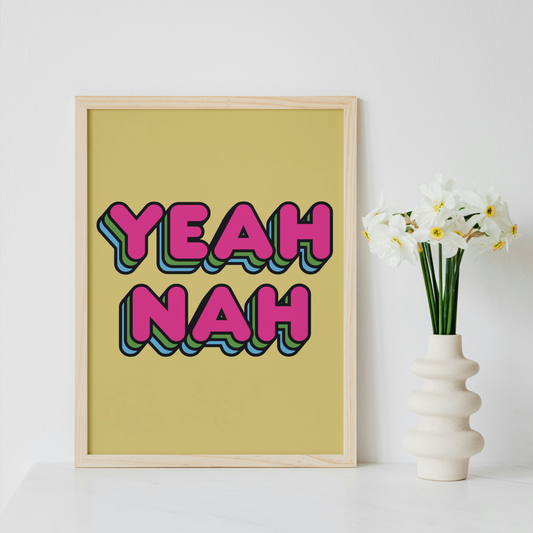 A framed typographic art print reading 'YEAH NAH' in pink bubble letters with blue and green shadowing, displayed on a white surface next to a white vase with white flowers against a white wall.