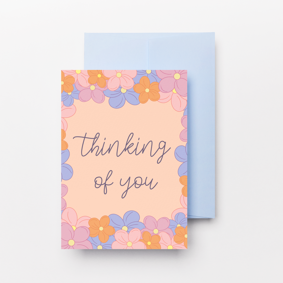 A greeting card with a peach background and a colorful floral border in pink, orange, and purple, with the words 'thinking of you' written in gray script. A light blue envelope is partially visible behind the card on a clean white background.