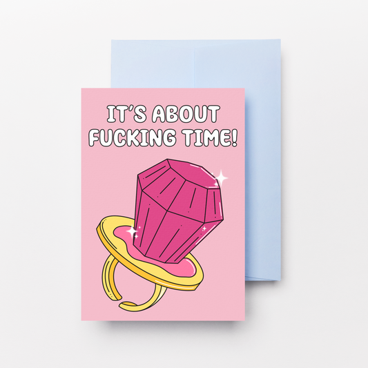 A pink greeting card with a cartoon pink ring pop on a yellow ring and the text 'IT'S ABOUT FUCKING TIME!' stacked on top of a light blue envelope against a light gray background.
