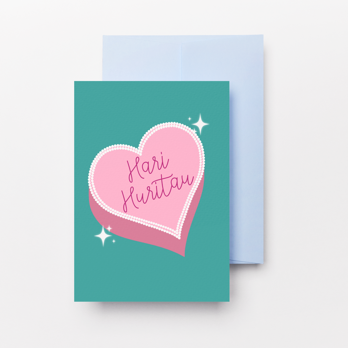 A teal greeting card with a pink heart graphic and the words 'Hari Huritau' in script, shown with a light blue envelope behind it on a clean white background.