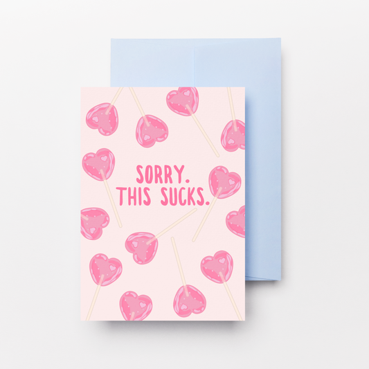 A greeting card with a light pink background, decorated with pink heart-shaped lollipops and the text 'SORRY, THIS SUCKS.' in pink lettering, shown with a light blue envelope behind it on a white background.
