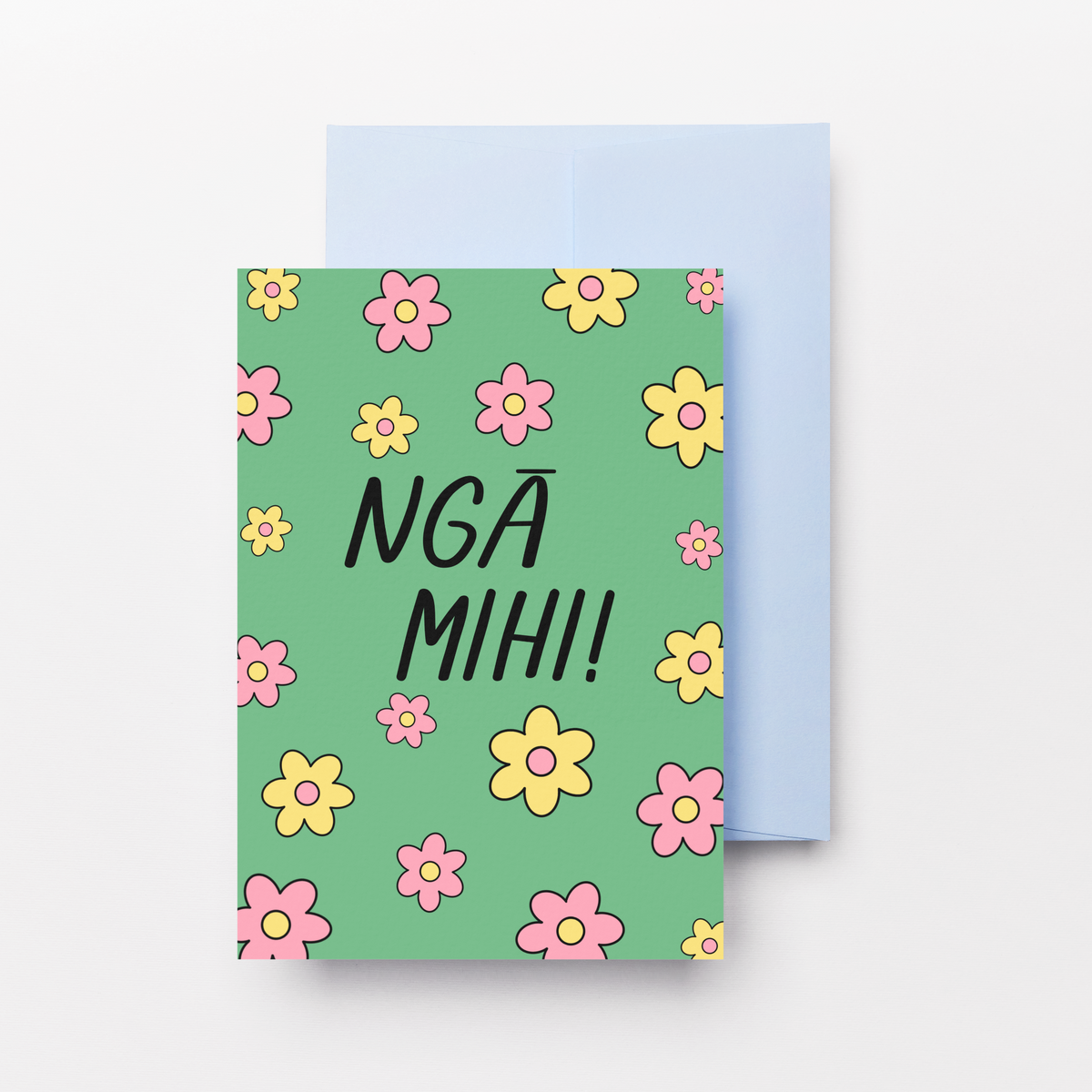 A greeting card with a mint green cover decorated with pink and yellow flowers and the text 'NGA MIHI!' in black, shown with a light blue envelope behind it on a white background.