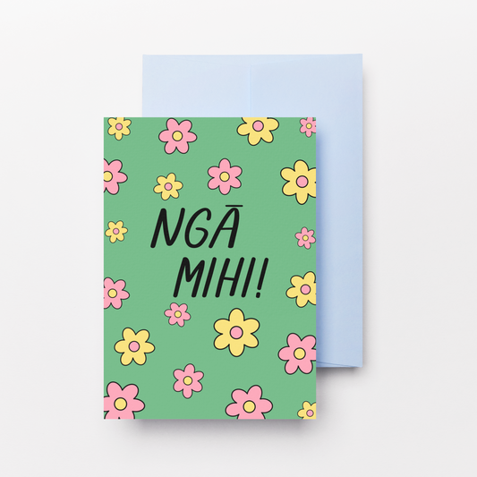A greeting card with a mint green cover decorated with pink and yellow flowers and the text 'NGA MIHI!' in black, shown with a light blue envelope behind it on a white background.