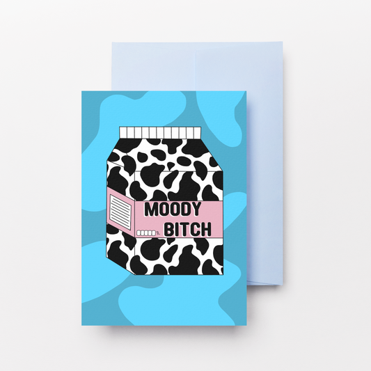 A greeting card with a light blue envelope behind it, featuring a graphic of a milk carton with a black and white cow print, a pink stripe, and the text 'MOODY BITCH' on a light blue abstract background.