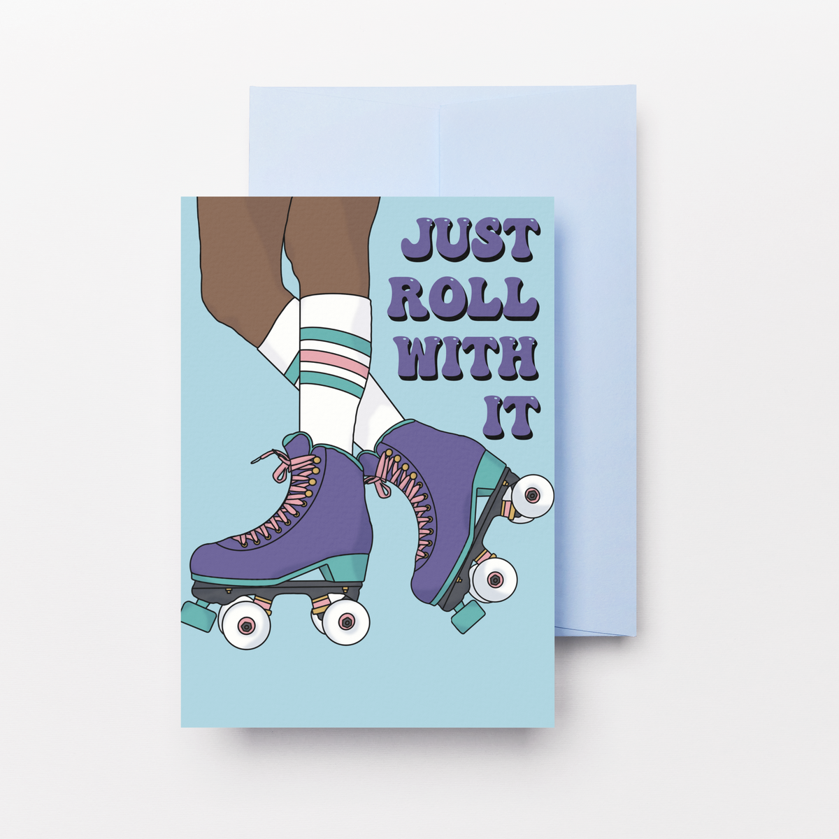 A rectangular greeting card with a light blue background and an illustration of a person roller skating, with the text 'JUST ROLL WITH IT' in dark purple. A light blue envelope is partially visible behind the card on a clean white background.