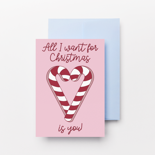 A pink Christmas greeting card with a candy cane illustration and the text 'All I want for Christmas is you!' shown in front of a light blue envelope on a white background.