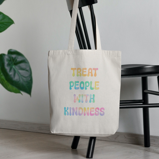 A cream canvas tote bag with the text 'TREAT PEOPLE WITH KINDNESS' in a pastel rainbow gradient, hanging from a black wooden chair in a minimalist room with a plant in the background.