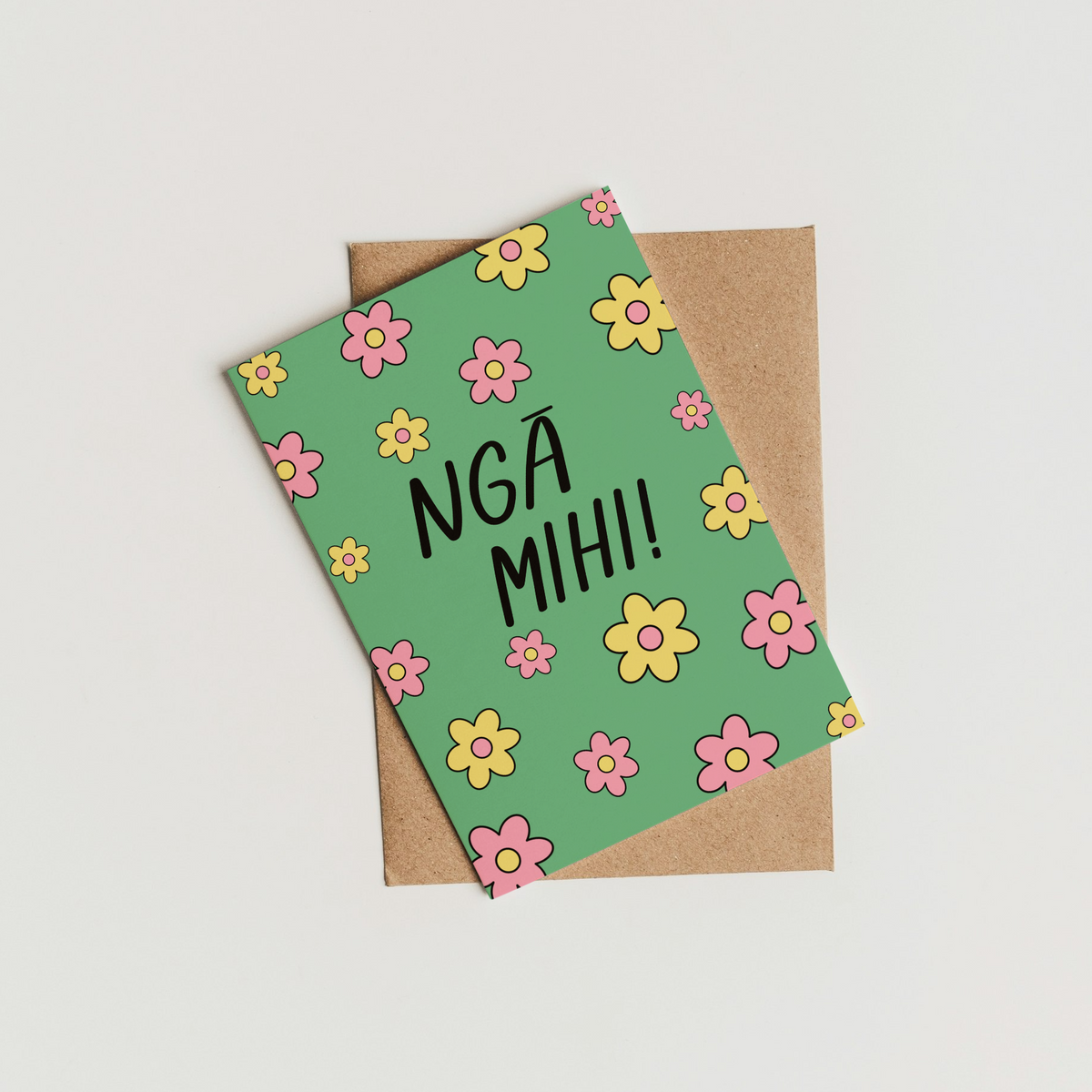 A green greeting card with colorful flower patterns and the text 'NGA MIH!' in black, resting on top of a brown kraft envelope against a plain white background.