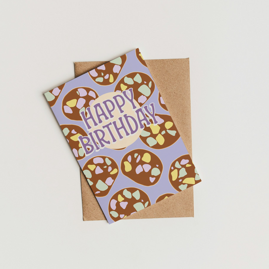 A square birthday card with a lolly cake pattern and the words 'HAPPY BIRTHDAY' in purple, placed on top of a brown kraft envelope against a plain off-white background.