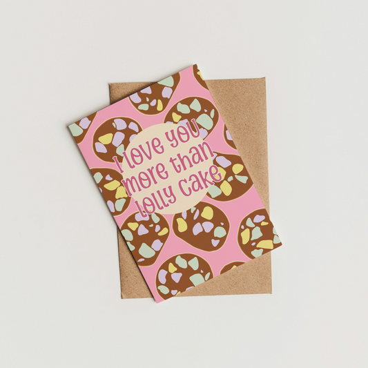 A pink greeting card with a repeating lolly cake pattern and the text 'love you more than lolly cake' in pink script, placed on top of a brown kraft envelope against a plain light background.