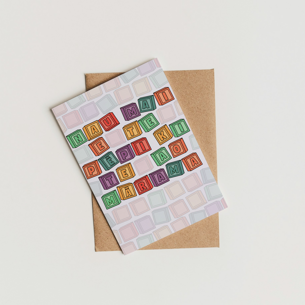 A square greeting card with a colorful grid pattern of small blocks and the words 'THANK YOU' repeated across it, placed on top of a brown kraft envelope against a plain off-white background.