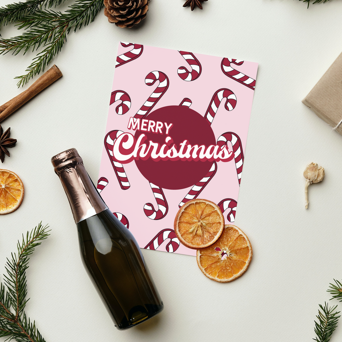 A festive flat-lay featuring a pink Christmas card with candy cane patterns and 'Merry Christmas' text, styled with a sparkling bottle, dried orange slices, pine branches, cinnamon sticks, and a wrapped gift on a white background.